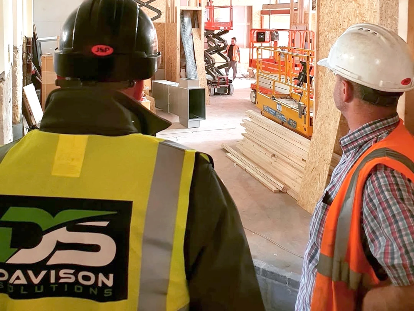 Two construction workers standing in a room with wooden framing, wearing safety gear and discussing the project.