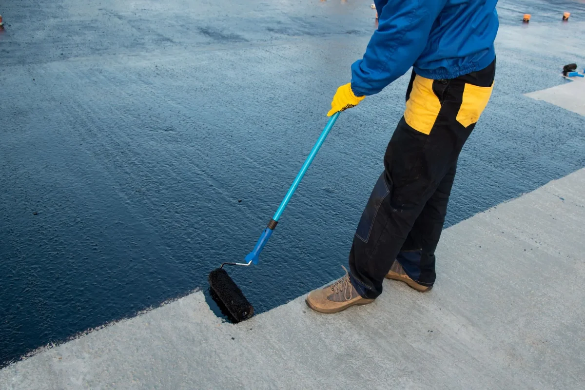 Worker applying black waterproof coating to concrete roof with roller.