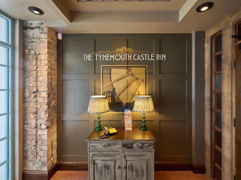A warm hospitality interior featuring an exposed brick pillar and wood-panelled walls with THE TYNEMOUTH CASTLE INN signage.