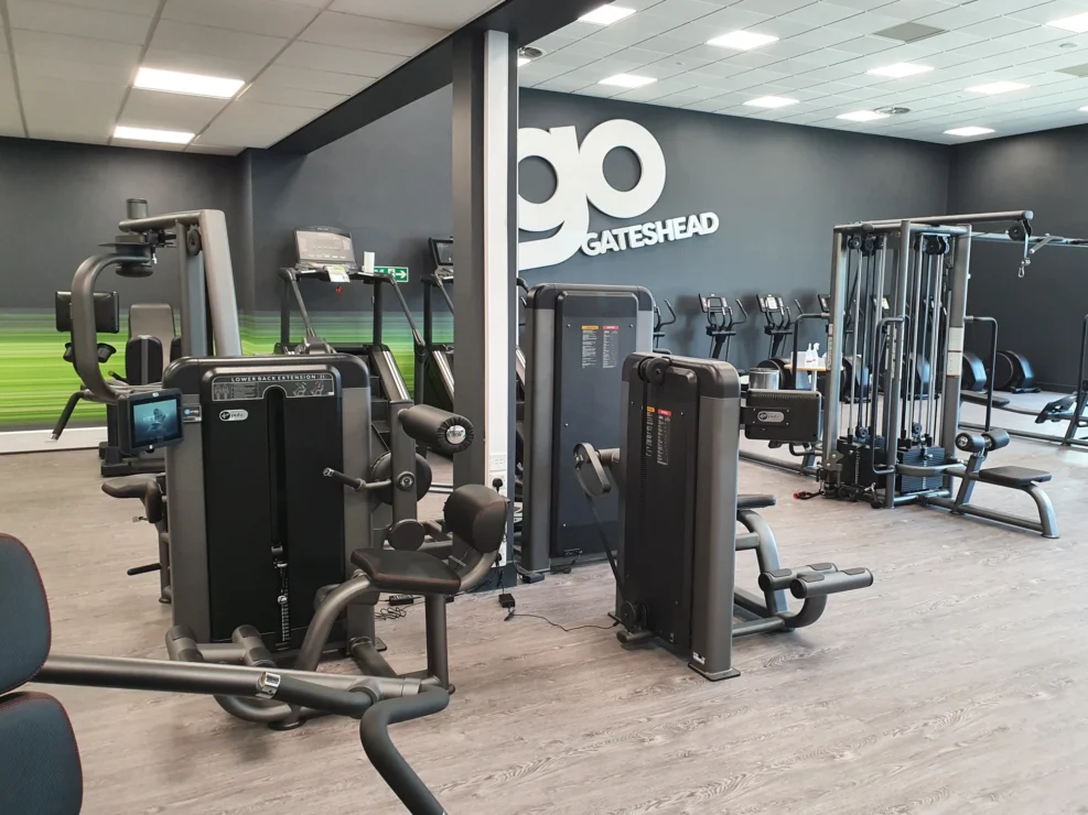 A modern commercial gym interior with grey flooring, black weight machines, and a prominent GO GATESHEAD logo on a black wall.