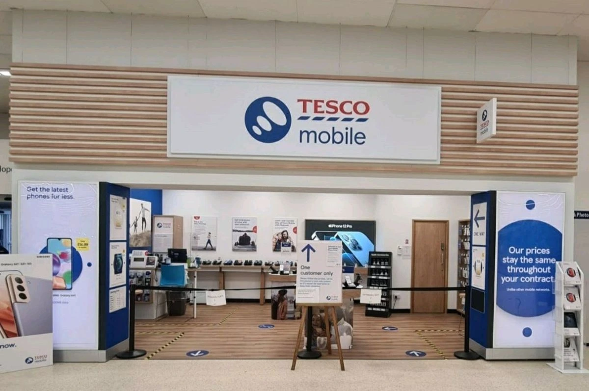 A Tesco Mobile store inside a supermarket with a black barrier blocking entry and a sign reading ‘One customer only’ on an easel.