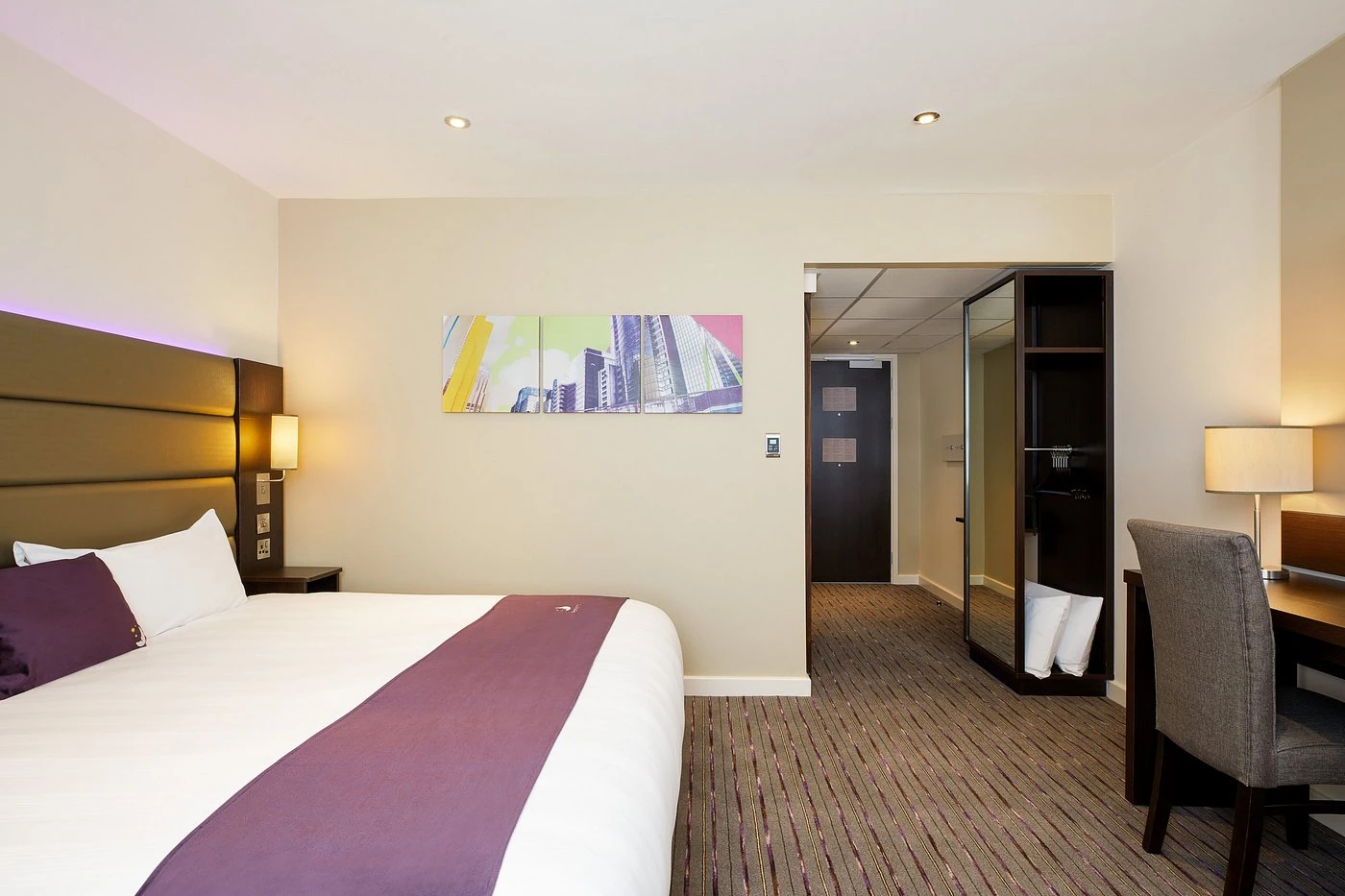 A modern hotel bedroom with neutral walls, a purple bed runner, and contemporary wall art.