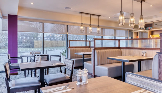 Modern restaurant dining area with grey leather booths, wooden tables, and a purple accent pillar.