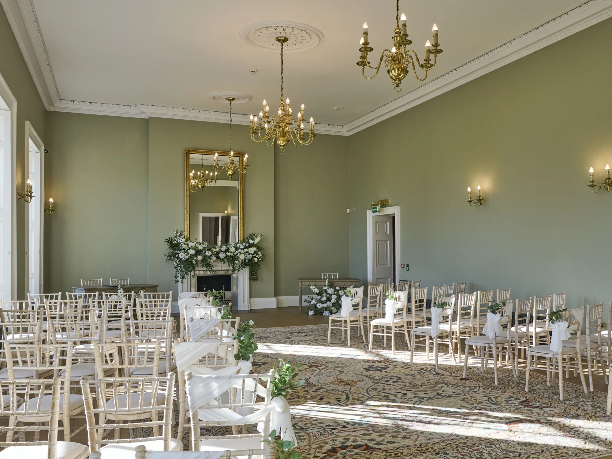 A grand events space set up for a wedding ceremony featuring sage green walls, high ceilings, and golden chandeliers.