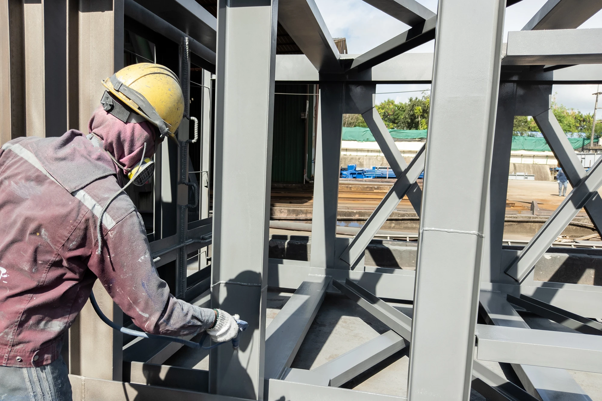 A worker wearing a yellow hard hat and protective gear uses a spray gun to apply an intumescent coating to a large industrial steel structure.