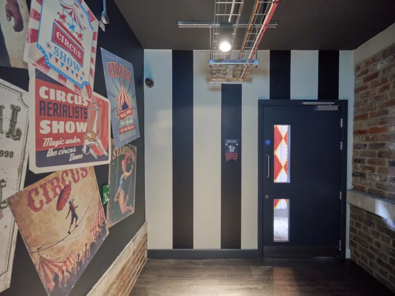 Interior hallway with black and white vertical striped walls, vintage circus show posters, and a black exit door next to an exposed brick wall.