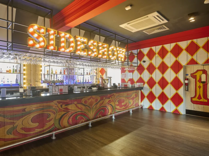 A circus-themed Sideshow bar with a large illuminated marquee sign, custom painted bar front, and a red and yellow diamond pattern wall.