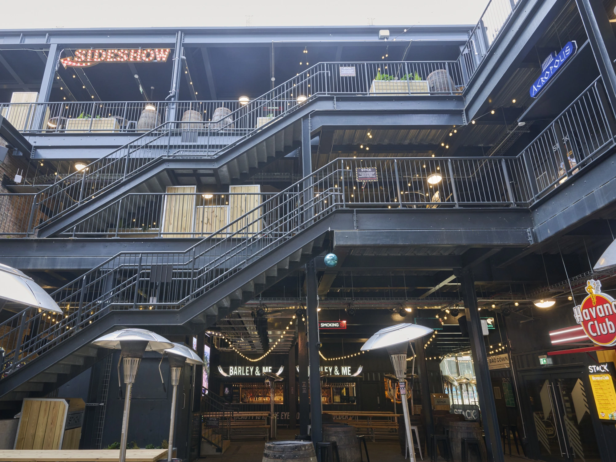 Multi-level outdoor entertainment courtyard showing black steel staircases, overhead balconies, patio heaters, and various bar signs.