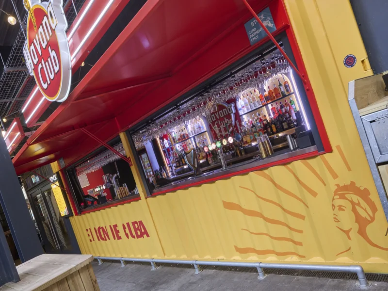 A vibrant yellow and red Havana Club bar container with a service hatch, illuminated bottle display, and overhead glassware racks.