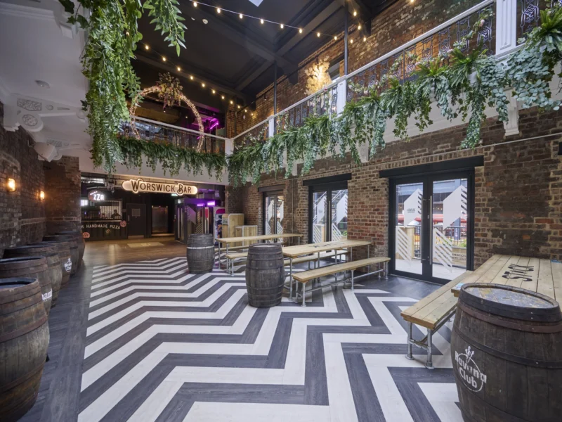 Indoor courtyard of Worswick Chambers featuring a chevron-patterned floor, wooden tables, beer barrels, exposed brick walls, and hanging green foliage.